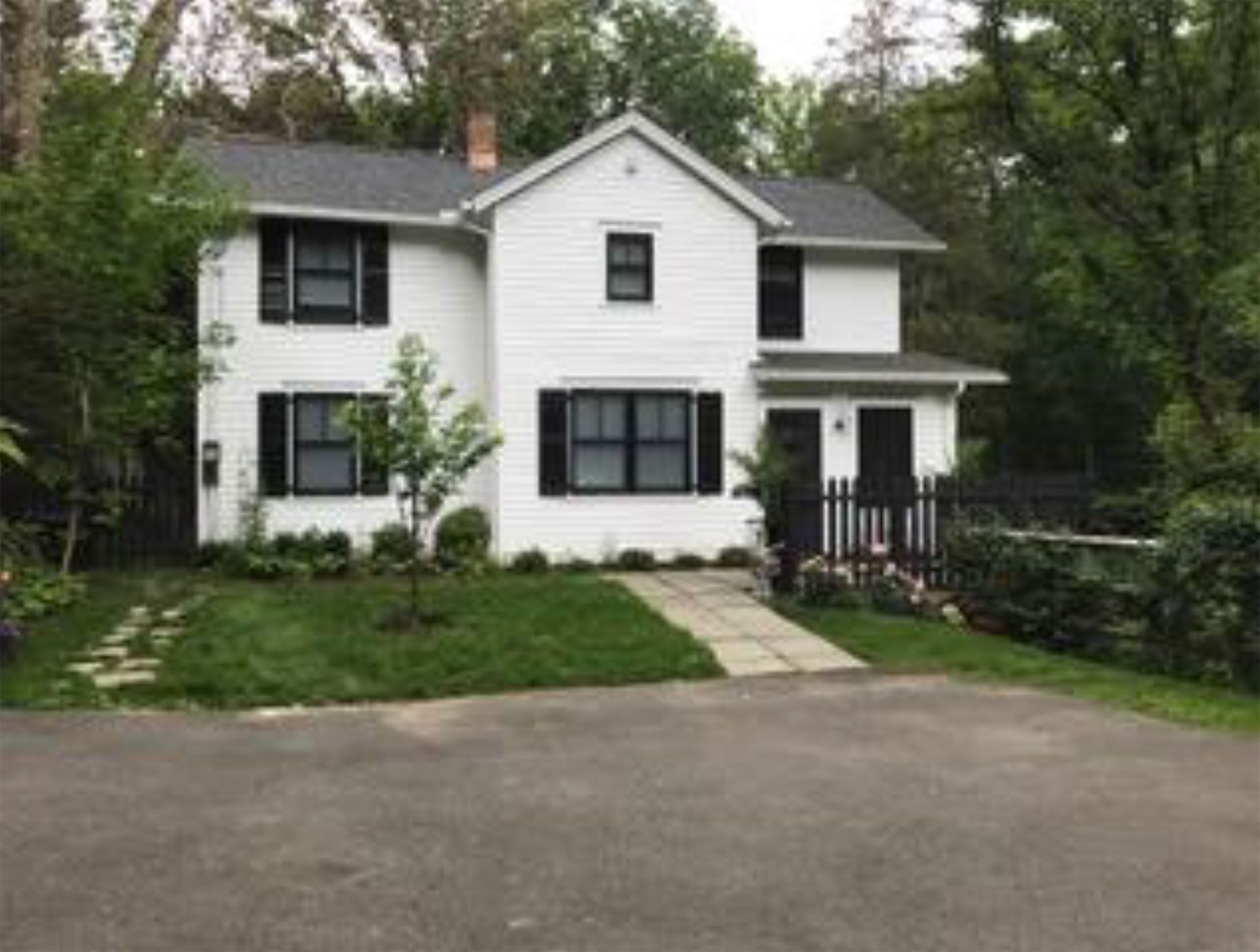 White two-story house with black shutters, surrounded by trees and grass, on a paved driveway.