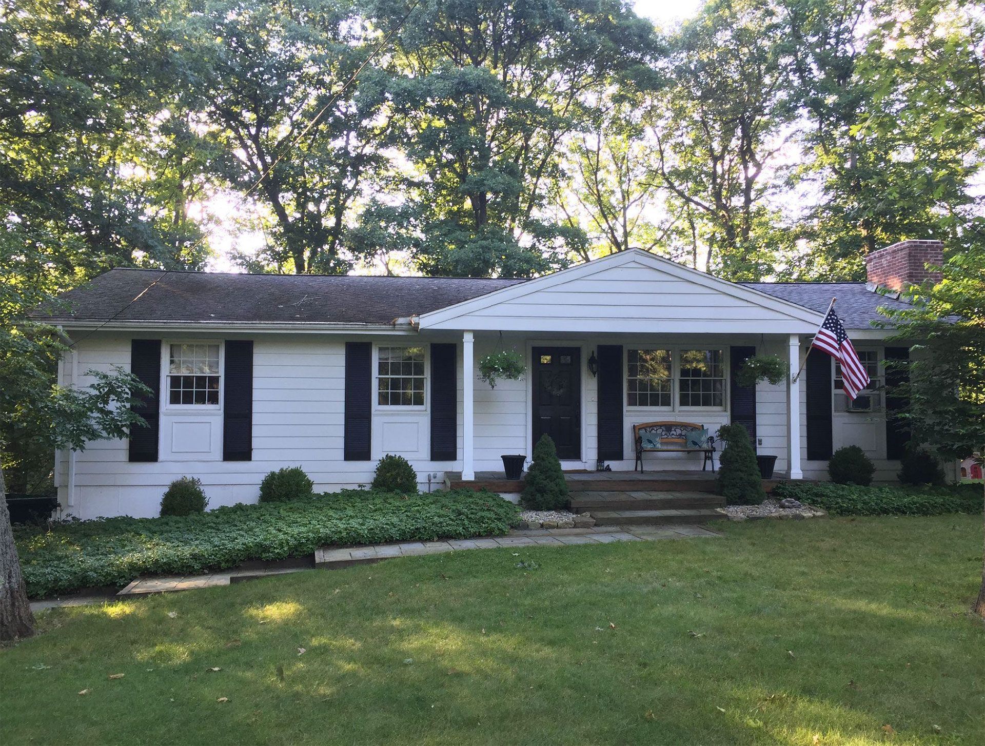 White ranch-style house with black shutters, green lawn, trees, and an American flag.