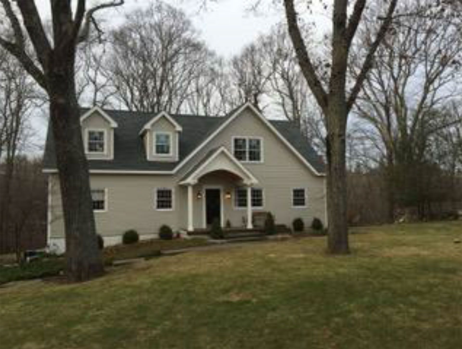 Beige two-story house with a dark roof and a grassy yard. Trees frame the house on a cloudy day.