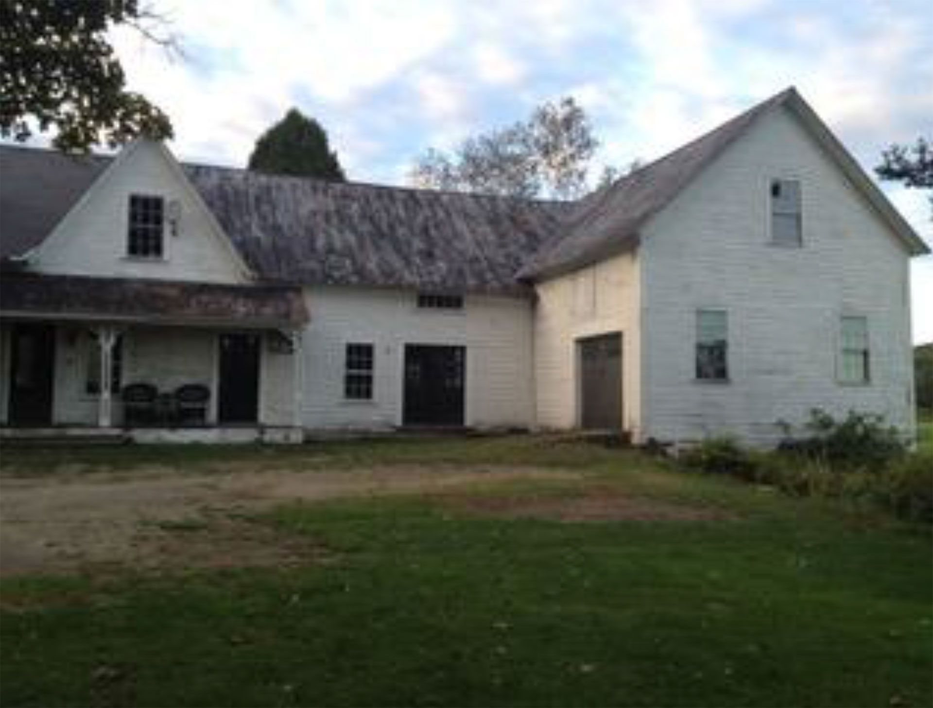 White farmhouse with porch, garage, and faded paint, set on a grassy lawn under a cloudy sky.