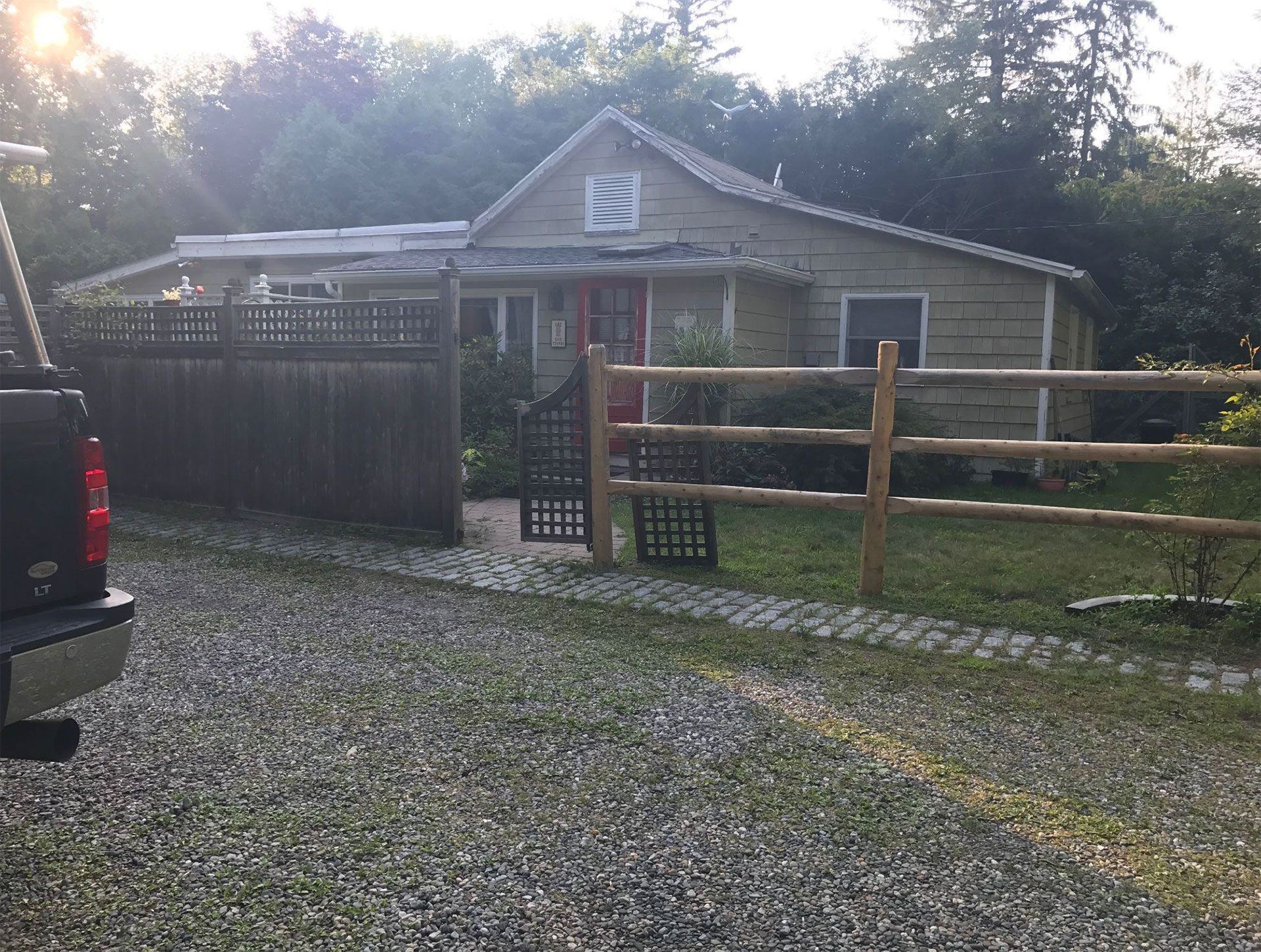 A gravel driveway leads to a one-story house with a wooden fence and a truck parked on the left.