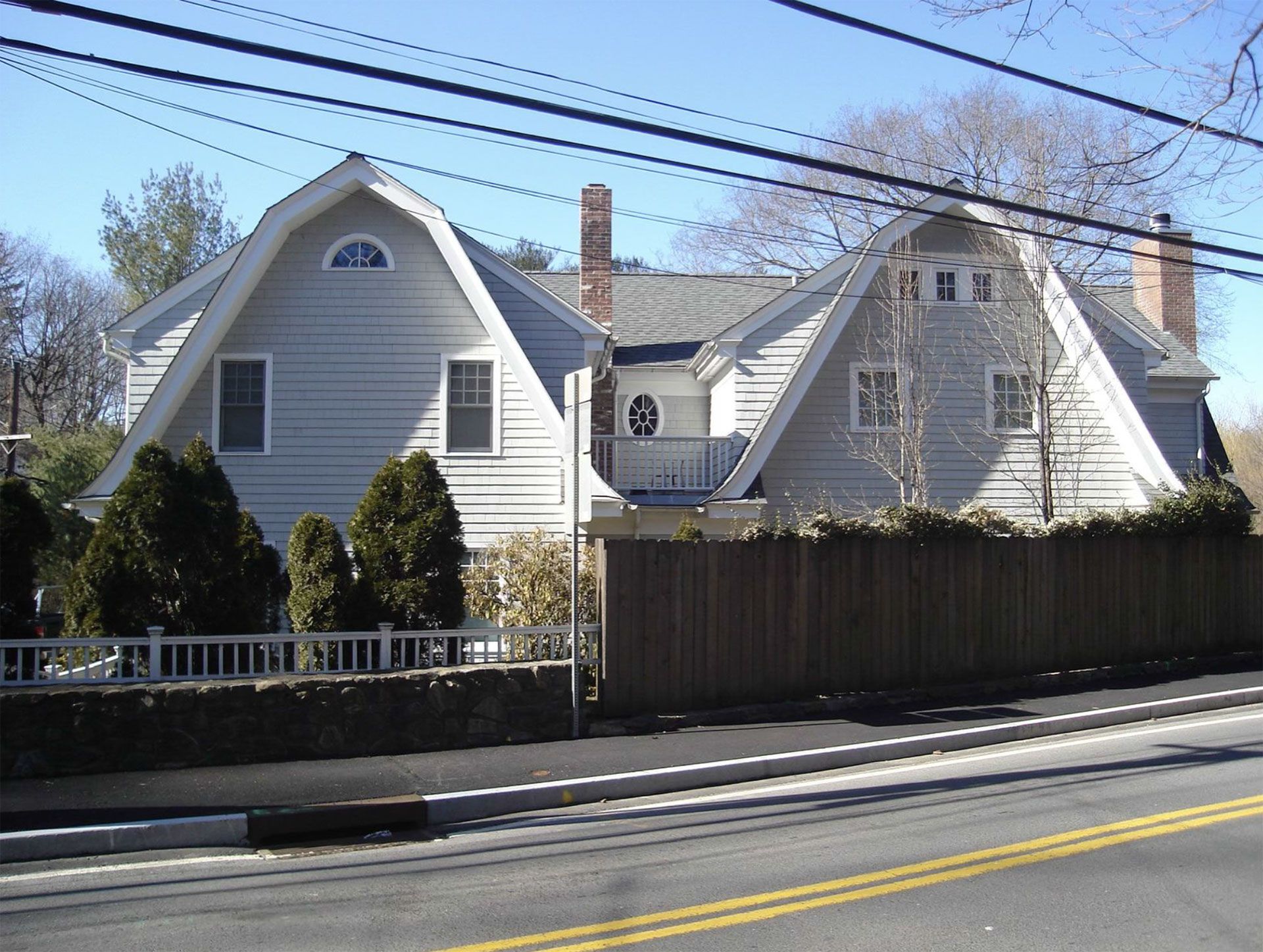Two-story light gray house with two angled roof sections, a chimney, and a wooden fence along a road.