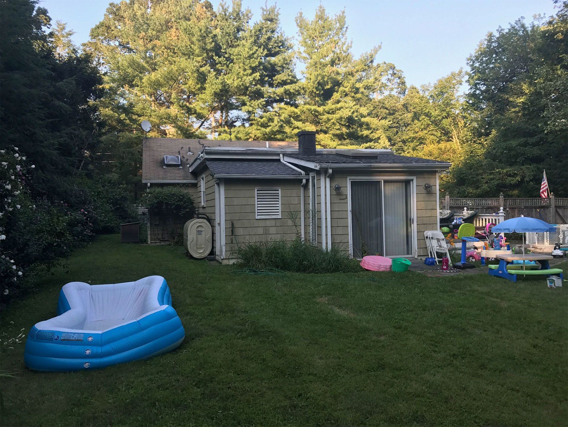 Backyard scene: Inflatable pool, small house, kids' toys on grass. Trees in background, sunny day.