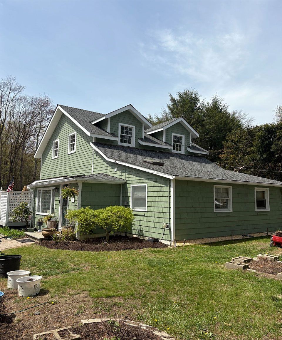 Green house with white trim, gray roof, and two dormers, set in a yard.