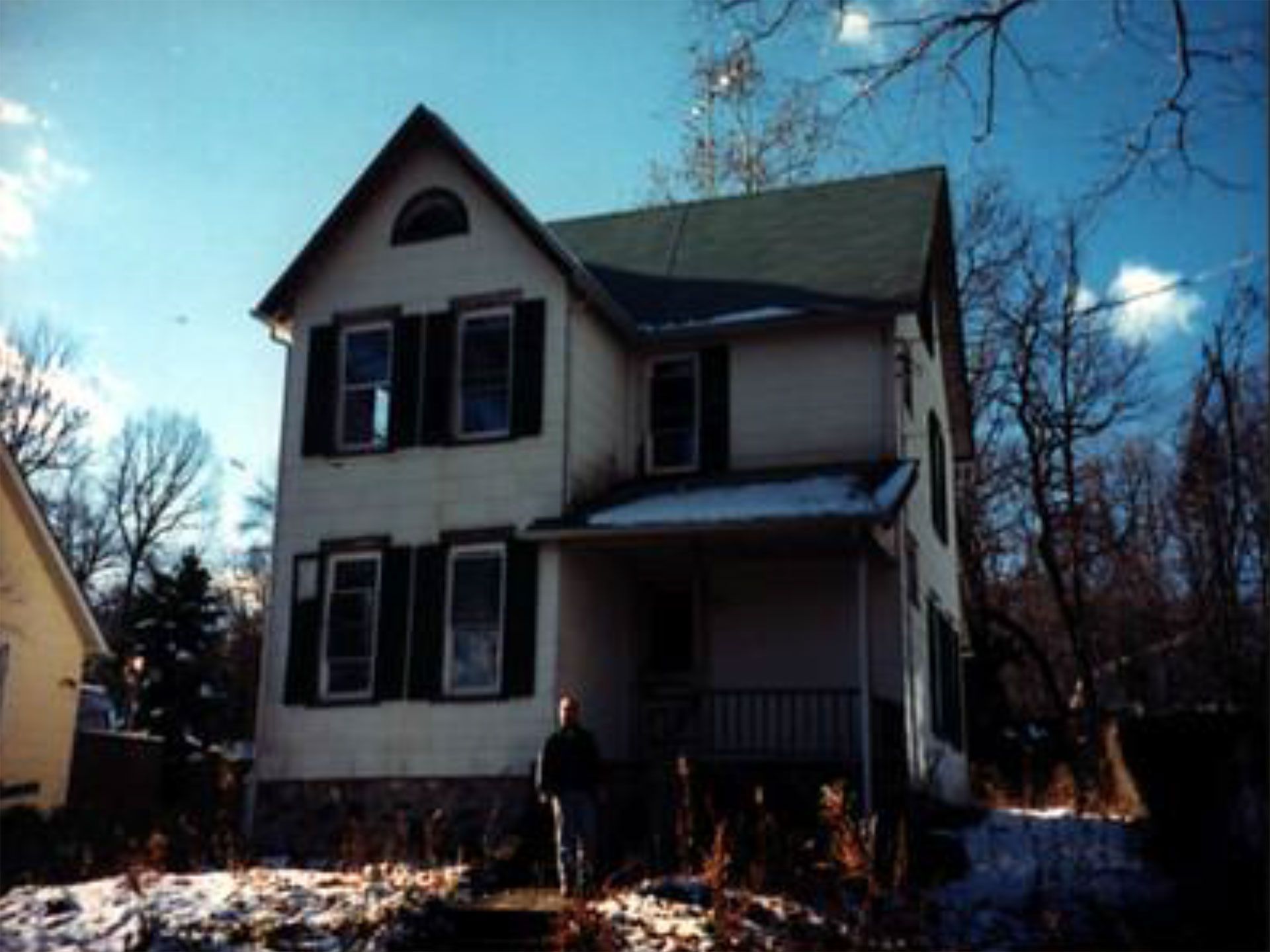Two-story house with green roof, black shutters, and porch. Man stands on steps in front of the house in winter.