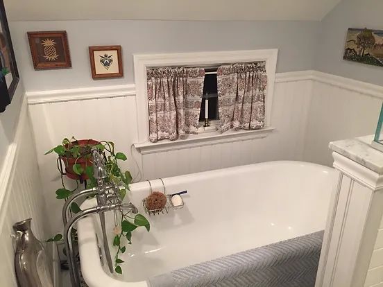 A vintage bathtub in a bathroom with a small window and patterned curtain; white wainscoting and blue walls.