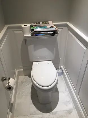 A white toilet in a small bathroom, with magazines stacked on the tank and a roll of toilet paper nearby.