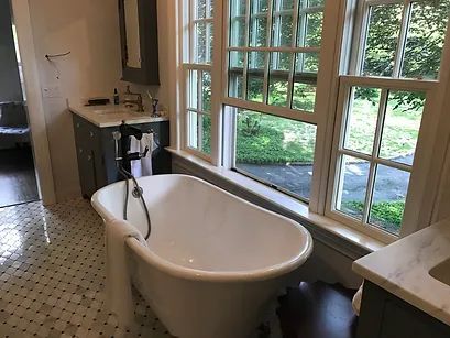 Bathroom with white clawfoot tub under a large window overlooking greenery. Marble countertops, tile floor.