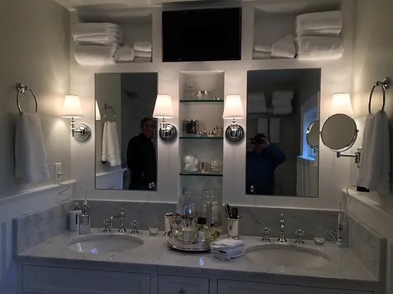 Elegant white bathroom with marble countertops, double sinks, mirrors, and a mounted TV.
