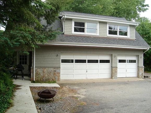 Two-car garage with small door and two windows above. Gray siding, white garage doors, stone accents.
