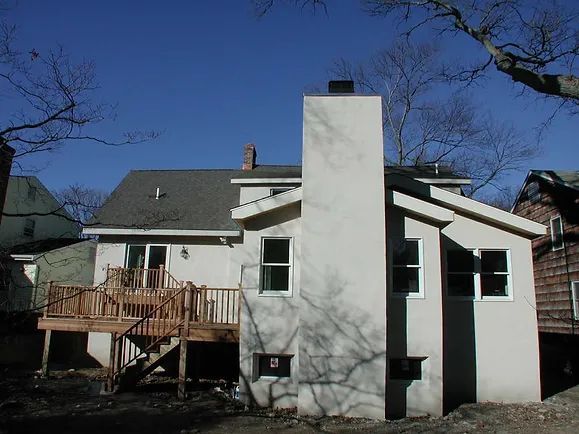 Back of a house with a wooden deck, chimney, and windows under a blue sky.