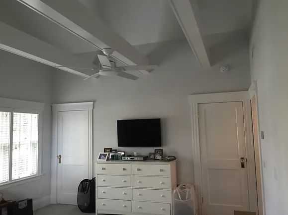 Bedroom with white dresser, TV, doors, windows, ceiling fan, and painted ceiling beams.