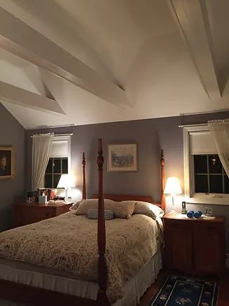 Bedroom with a four-poster bed, nightstands, windows, and exposed ceiling beams. Soft lighting and blue-grey walls.