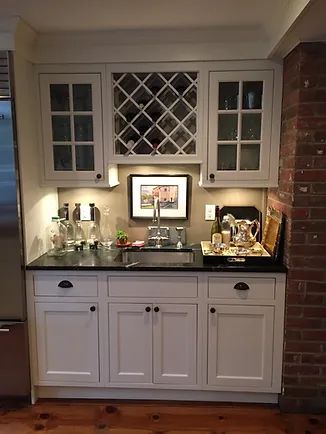 White bar cabinet with sink, wine rack, and glass-fronted cabinets against a brick wall.