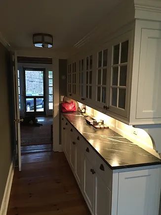 Narrow kitchen with white cabinets, dark countertop, and view through a doorway to the outside.