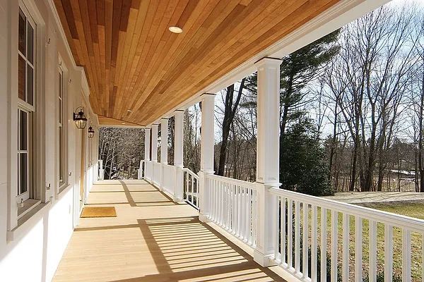 A white porch with columns, wood ceiling and railing, looking out to trees and a yard on a sunny day.
