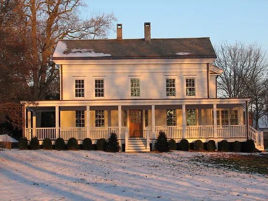 White two-story house with wraparound porch in a snow-covered field.