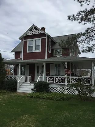 Two-story maroon house with white porch, trim, and decorative details; green lawn under cloudy sky.