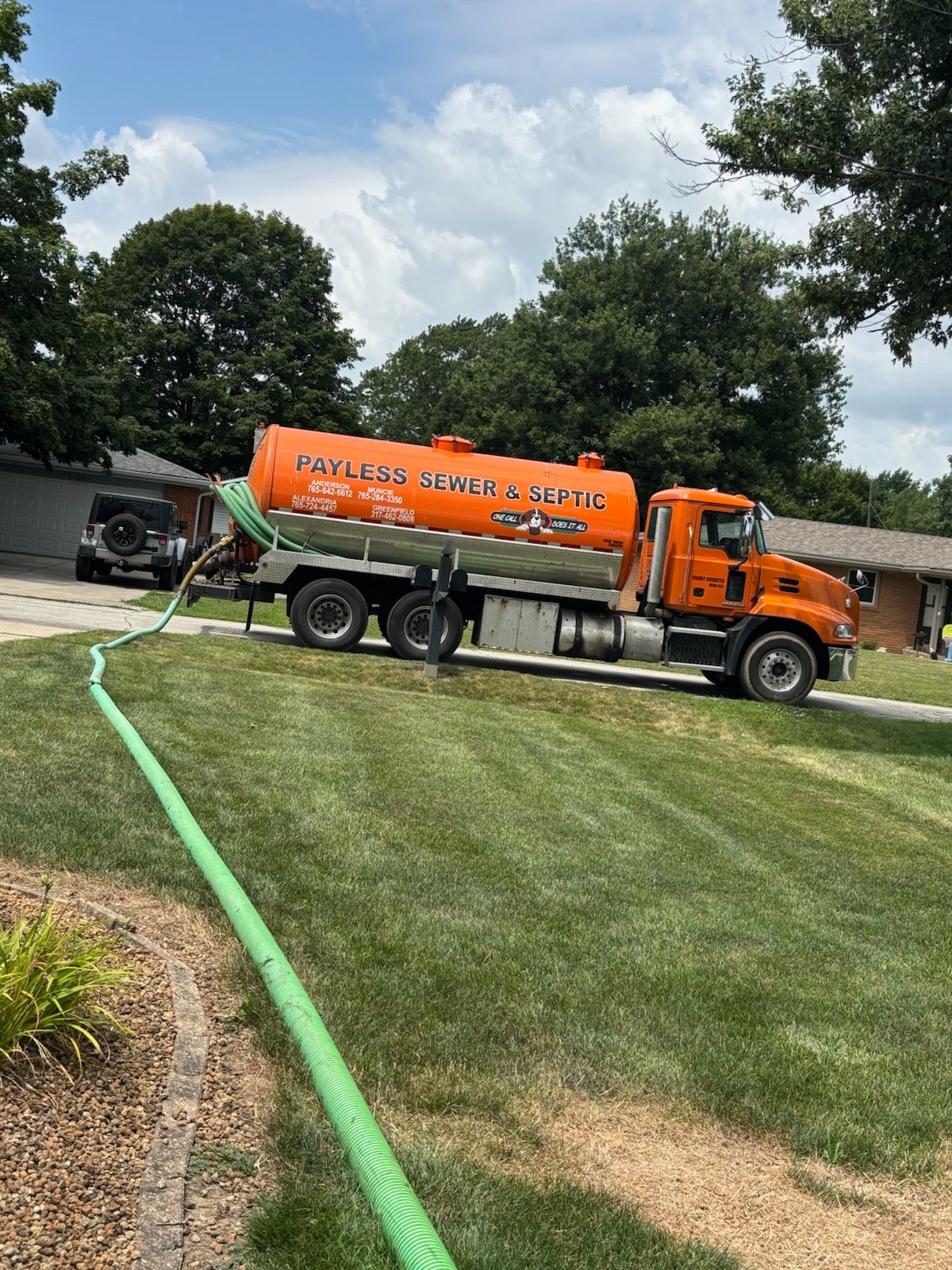 Orange tanker truck pumping green liquid on a grassy lawn near a driveway under a cloudy sky