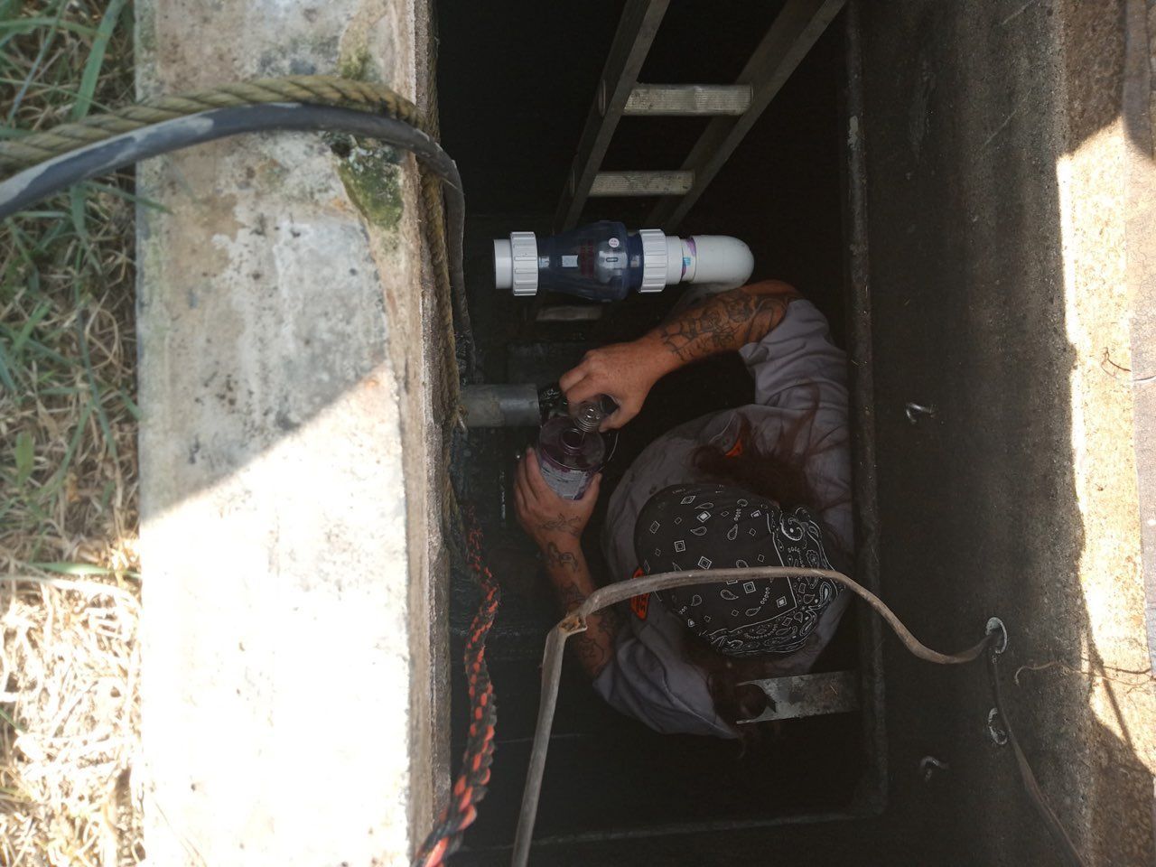Worker crouching in a narrow utility shaft, handling pipes and cables in a dim concrete space