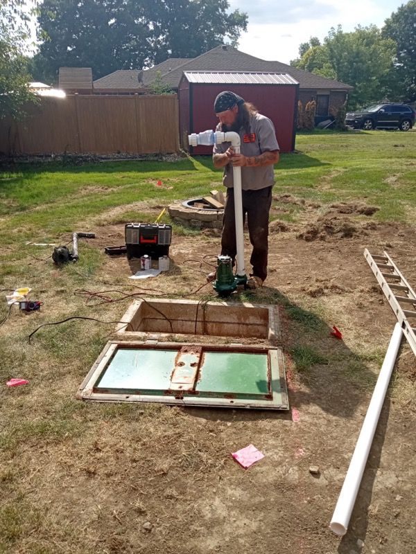 Person standing by an open underground hatch in a backyard, holding a tool near a green pipe and construction materials.