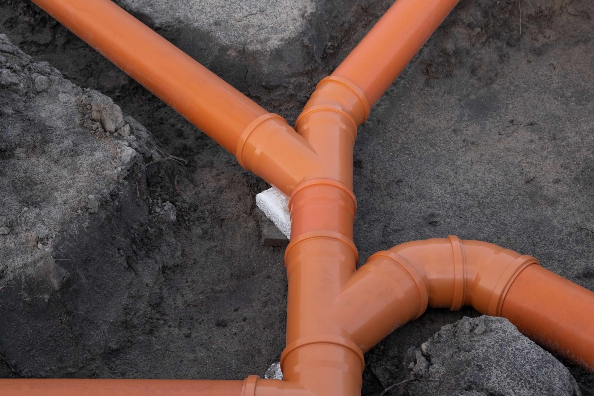 Orange plastic drainage pipes meeting at a Y-junction in dark soil and rocks
