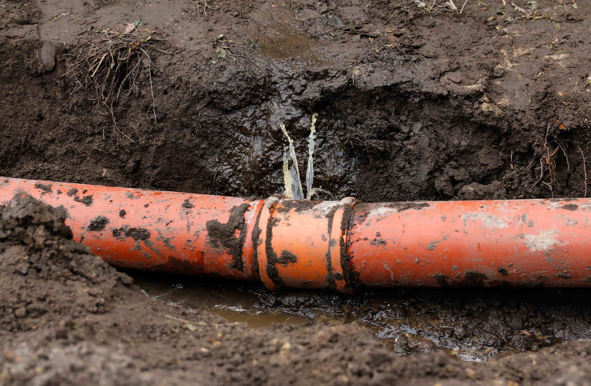 Orange pipeline leaking in muddy soil with water spraying from a crack