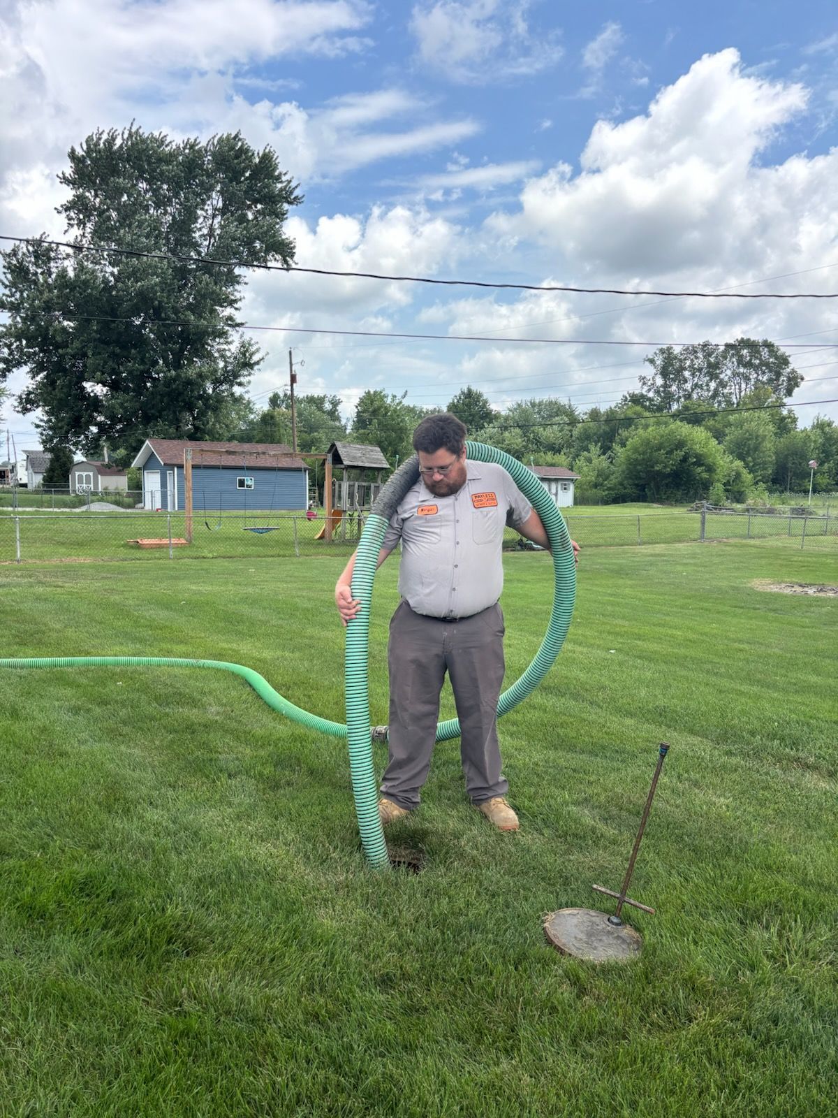 Man holding a green hose in a grassy yard under a cloudy sky, with a small shed in the background