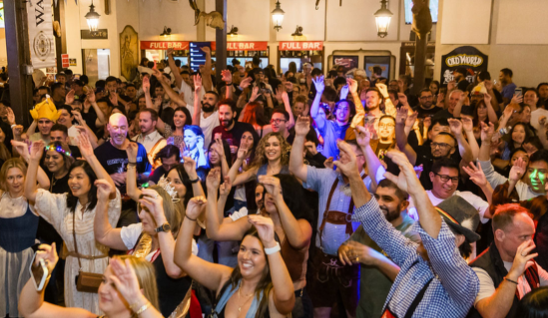 Large crowd of people with arms raised, celebrating in a dimly lit room, likely a pub or hall, with food and drink stalls visible.