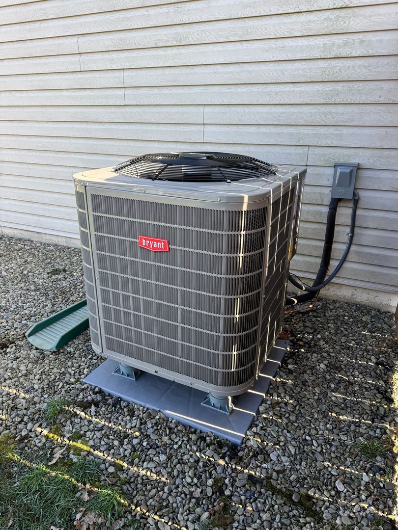 Outdoor air conditioner condenser unit beside a house wall on gravel, with a hose nearby.
