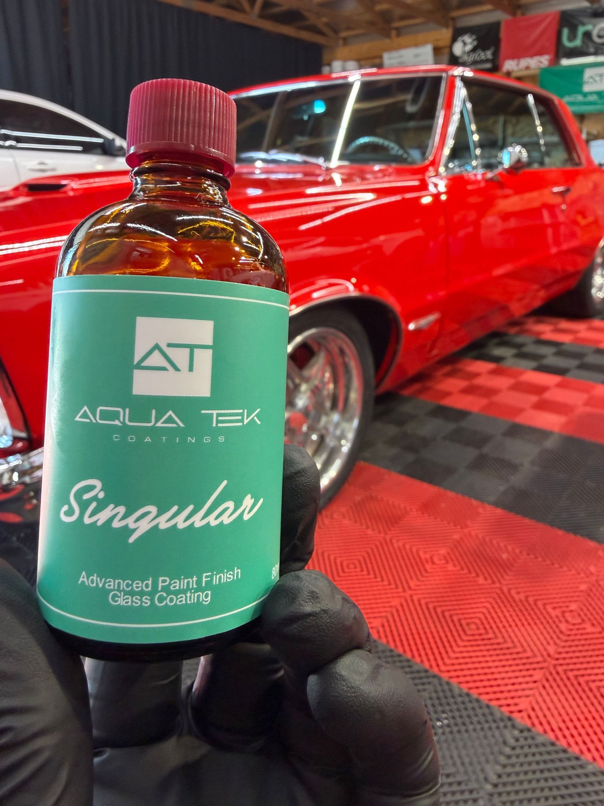 Hand holding an Aqua Tek Singular bottle in front of a red classic car display.
