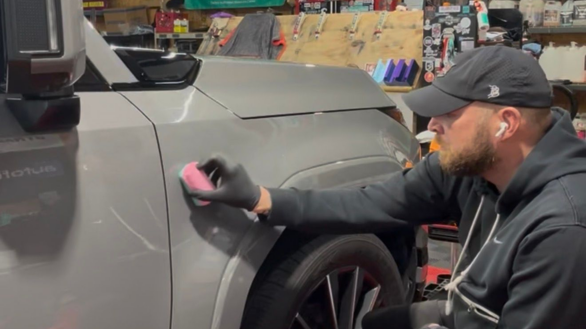 Person in a black cap and jacket cleaning a gray car wheel at an auto show.