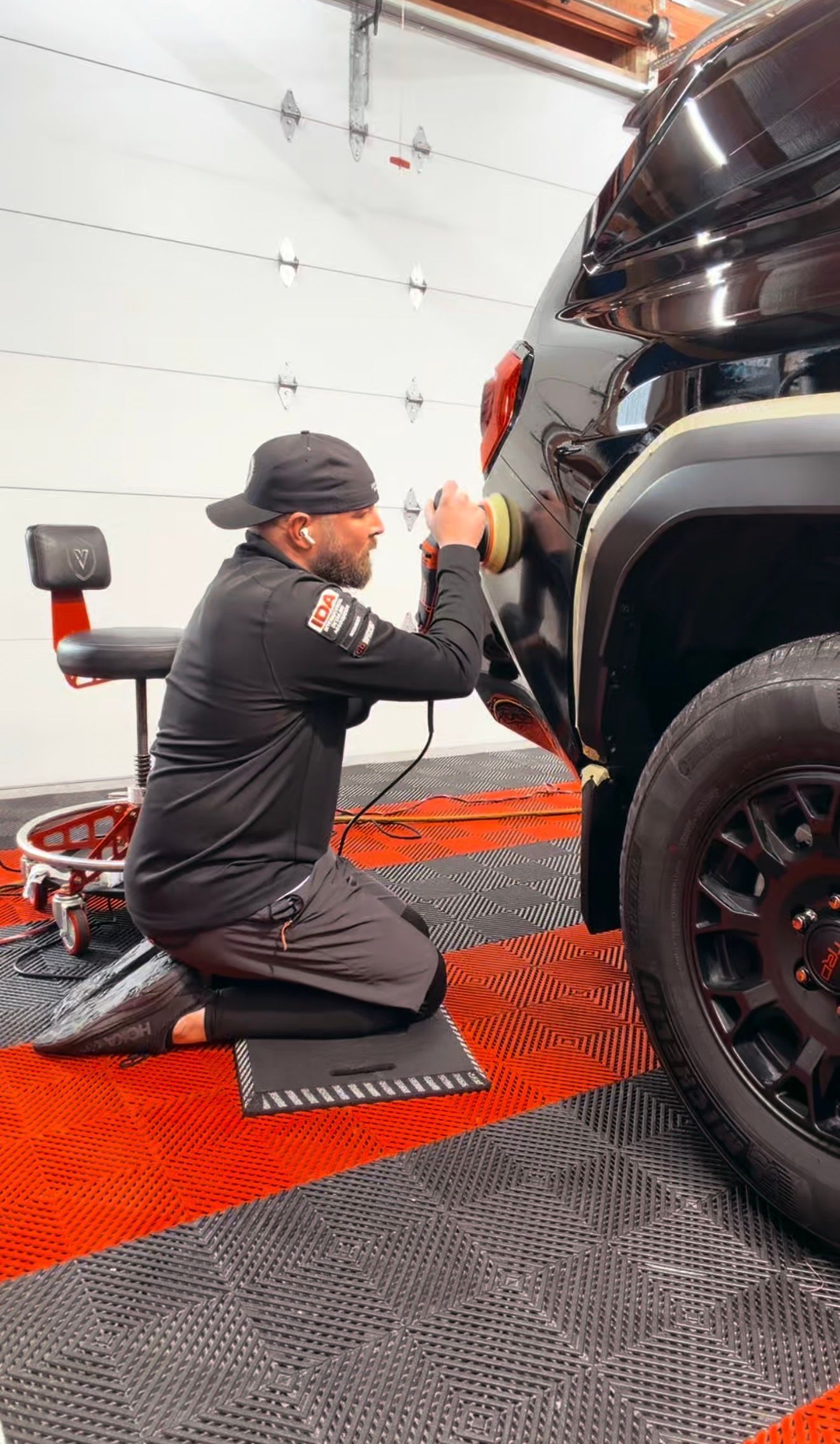 Mechanic kneels beside a black truck in a garage, inspecting the wheel area with a tool.