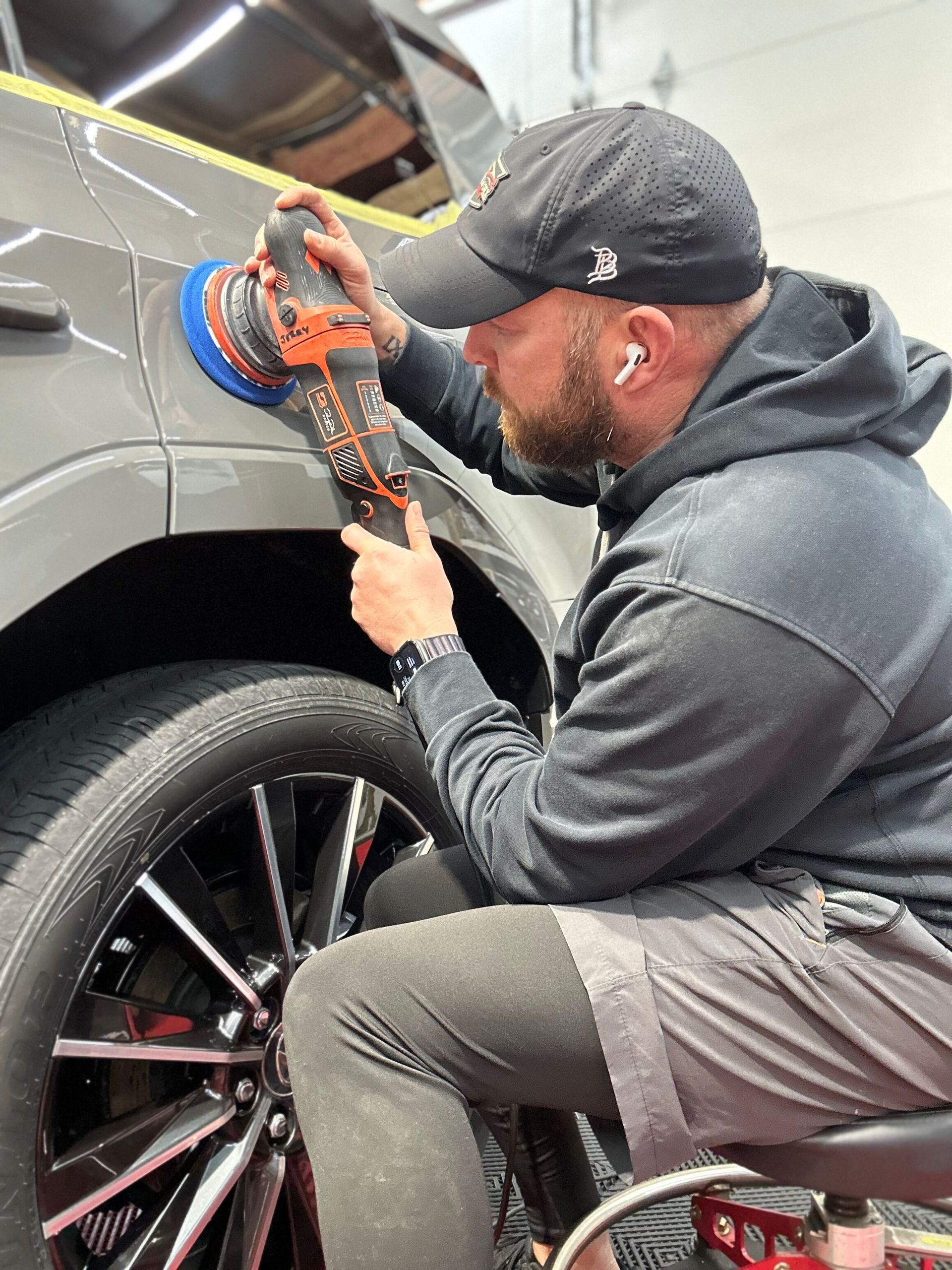 Mechanic polishing a gray car’s wheel arch with a handheld buffer in a garage