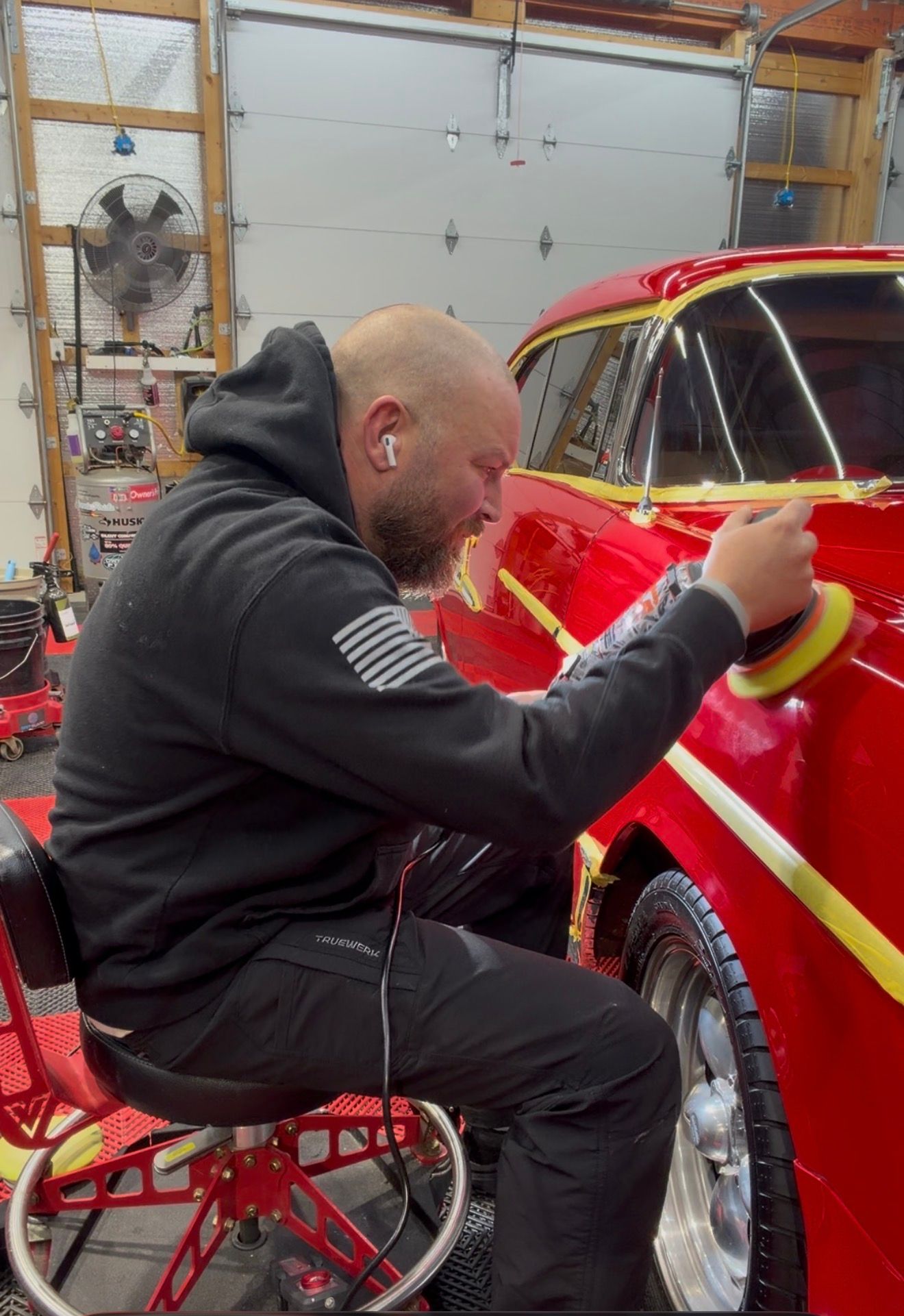 Person polishing a red car in a garage, seated on a low rolling stool.