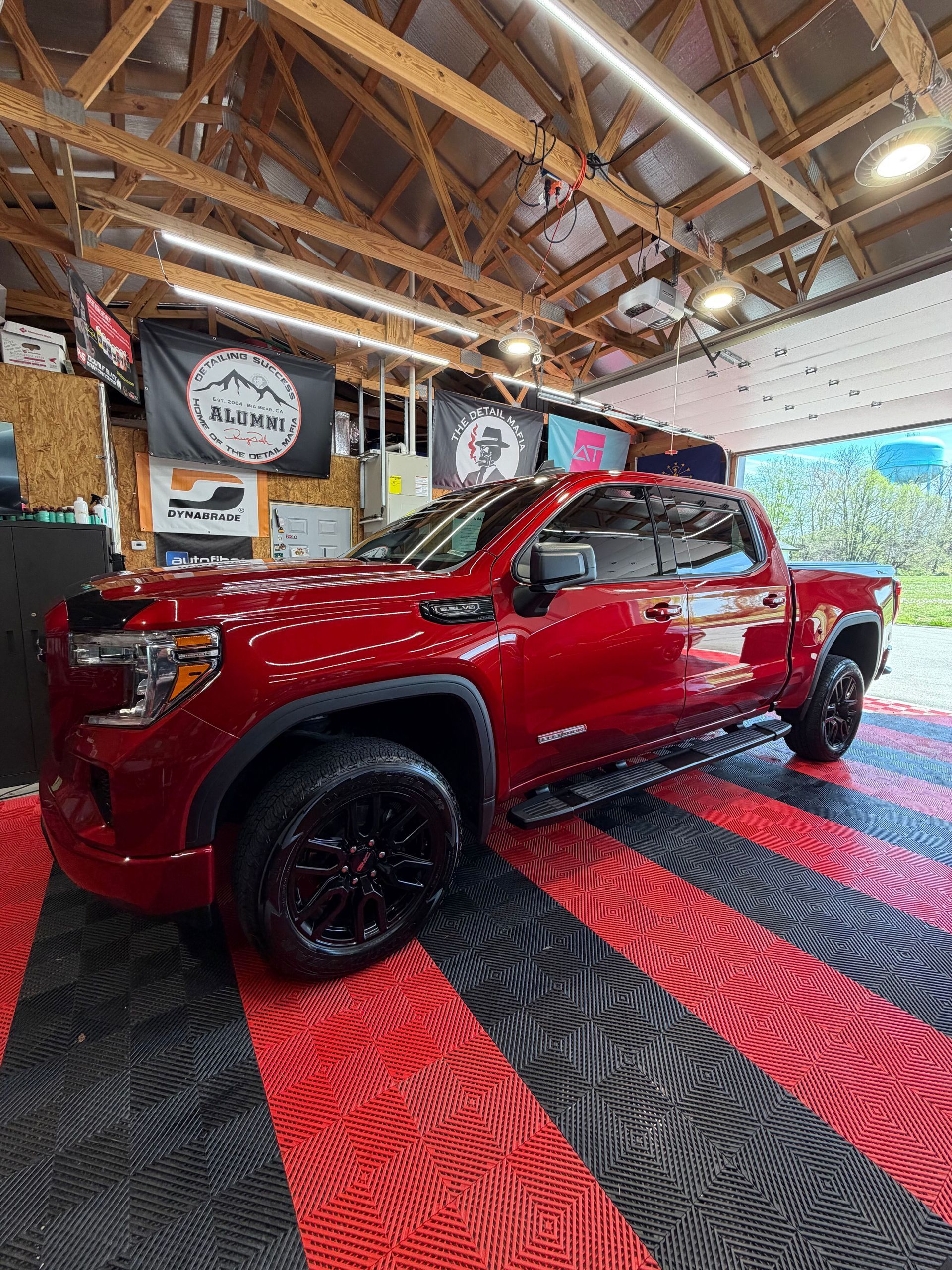 Red pickup truck in a workshop with red and black patterned flooring and exposed wooden rafters.