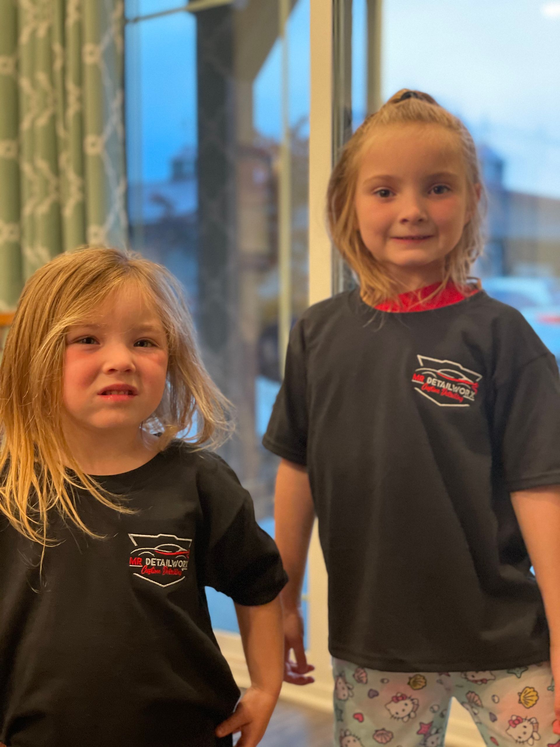 Two young girls wearing black t-shirts, standing near a window. One girl has a furrowed brow.