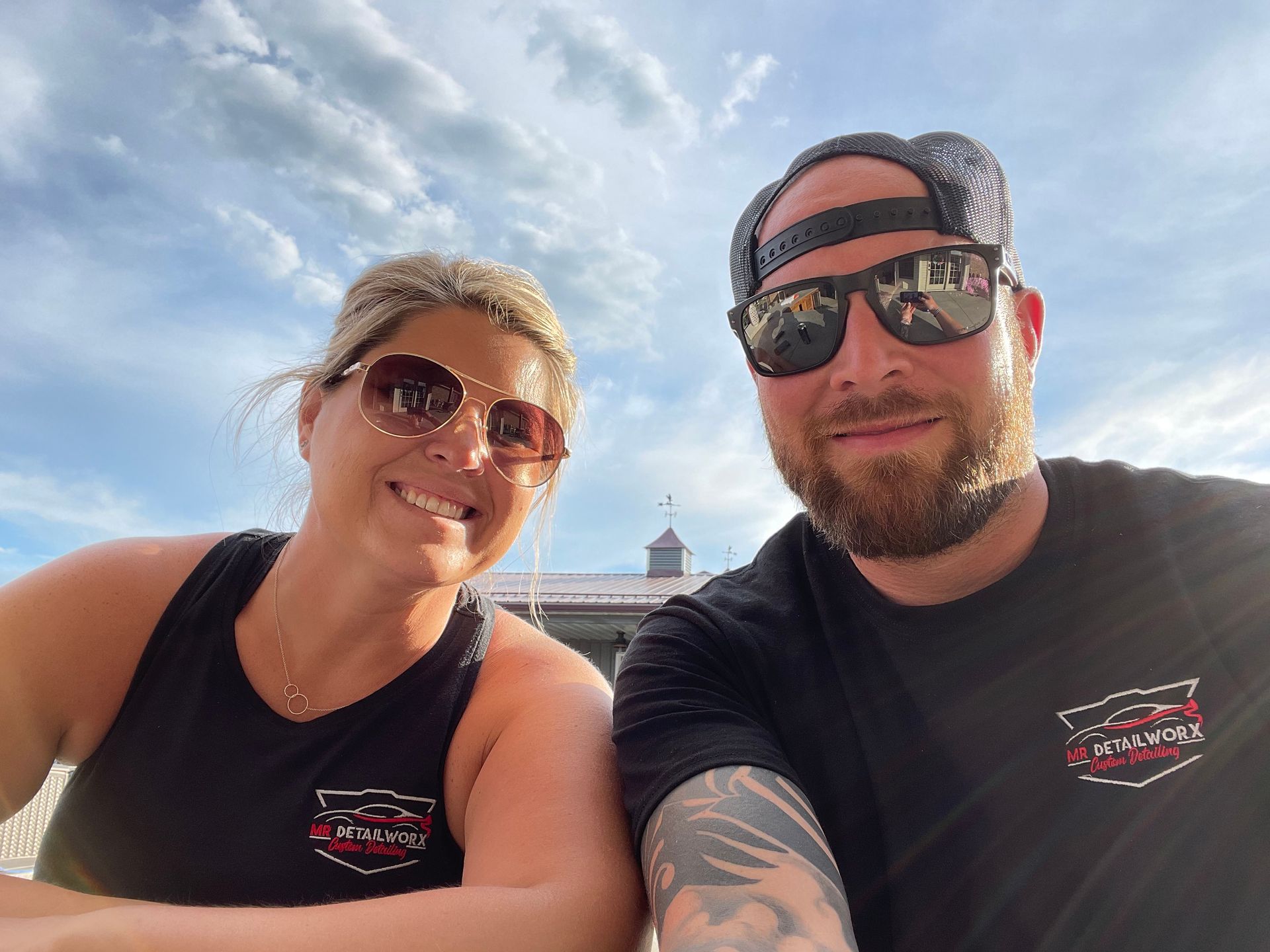 A woman and a man wearing sunglasses, smiling for a selfie outdoors. Blue sky, building in background.