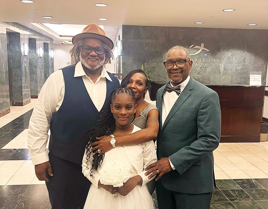 Four people pose together in a building lobby, including one person in a hat, a child, and two people in formal attire.