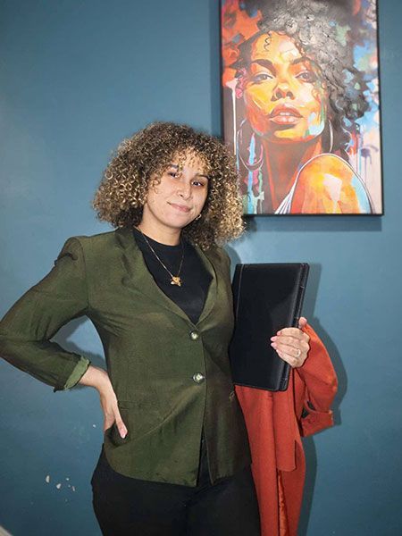 A person with curly hair wears a green blazer and holds a black folder, standing before a colorful portrait on a blue wall.