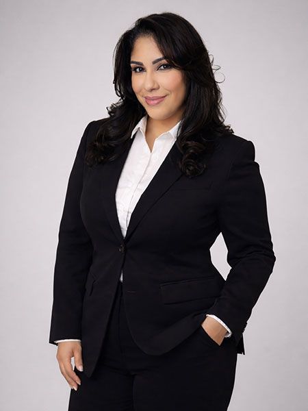 A smiling person in a dark suit and white shirt poses against a neutral gray backdrop, with one hand in a pocket.
