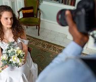 A photographer holds a camera to capture a portrait of a person in a white dress holding a bouquet in a vintage room.