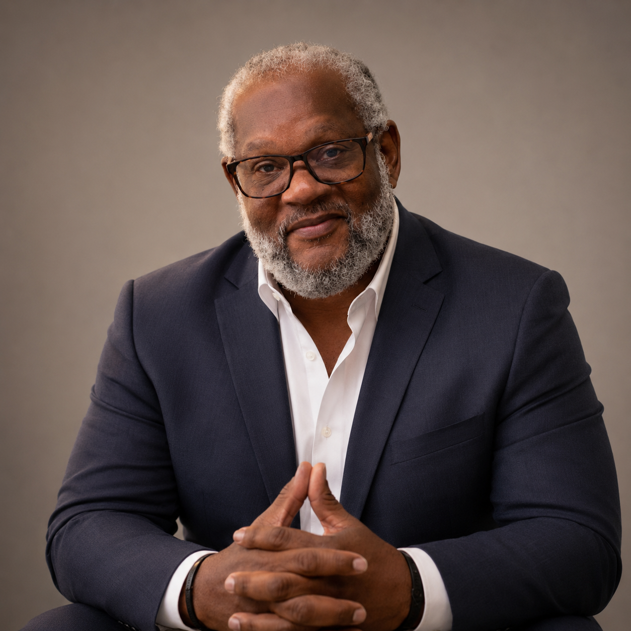 A person with grey locs and glasses wears a light blue button-up shirt against a plain grey background.