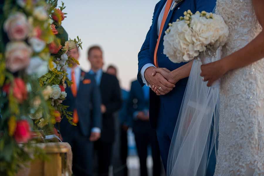 A couple holding hands at an outdoor wedding, the person on the right holding a white bouquet with a lace veil.
