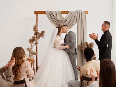 A bride and groom kiss under a draped wedding arch while guests clap in a minimalist, sunlit space.