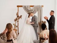 A bride and groom kiss under a draped wedding arch while guests clap in a minimalist, sunlit space.