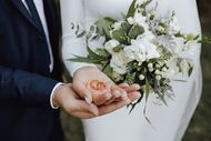 A person in a white gown and another in a dark suit hold a pair of gold wedding rings in their cupped hands with a bouquet.