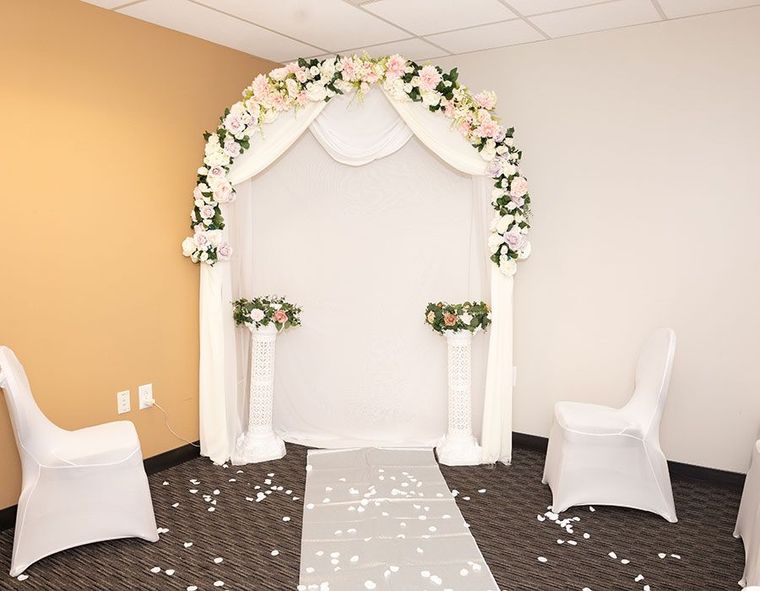 A floral wedding arch with white fabric drapery stands behind two white pedestals, flanked by two chairs on a gray runner.