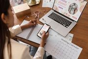 A person works at a desk, checking a phone while writing in a notebook near a laptop displaying wedding planner content.