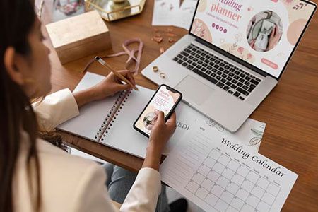 A person works at a desk, checking a phone while writing in a notebook near a laptop displaying wedding planner content.